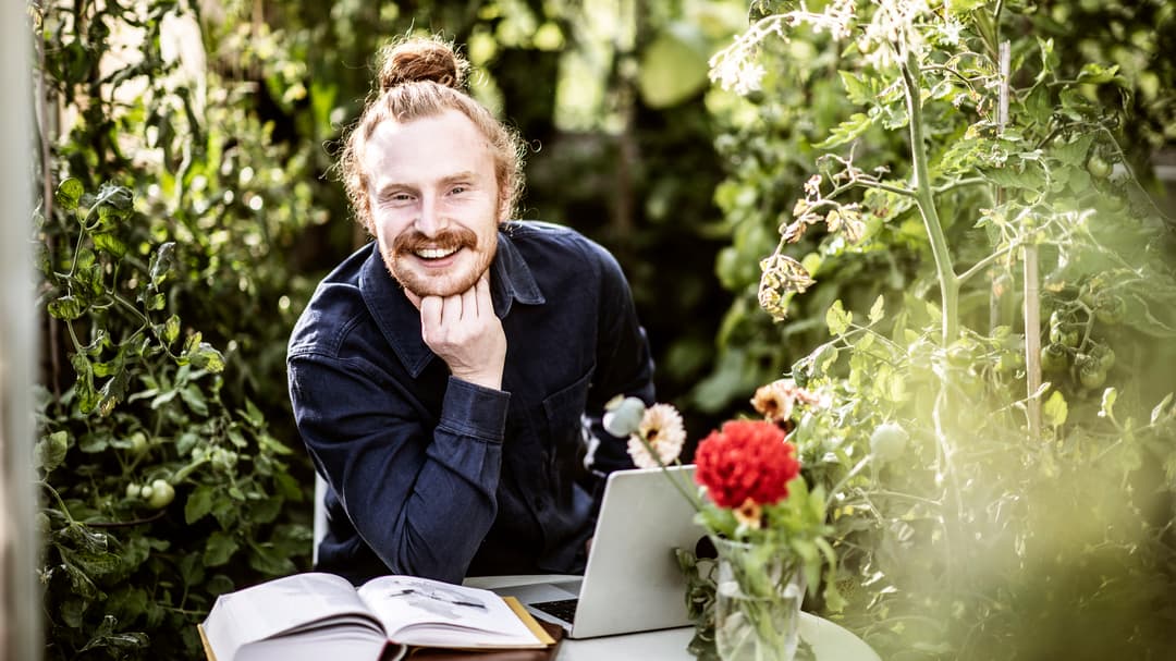 Manlig student sitter i  växthus och pluggar med laptop och böcker. 