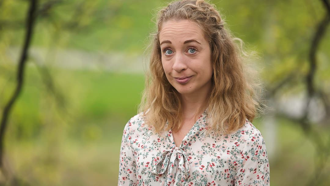 Kvinna med lockigt axellågt hår och blommig blus står utomhus med grönskande träd runt om kring sig.