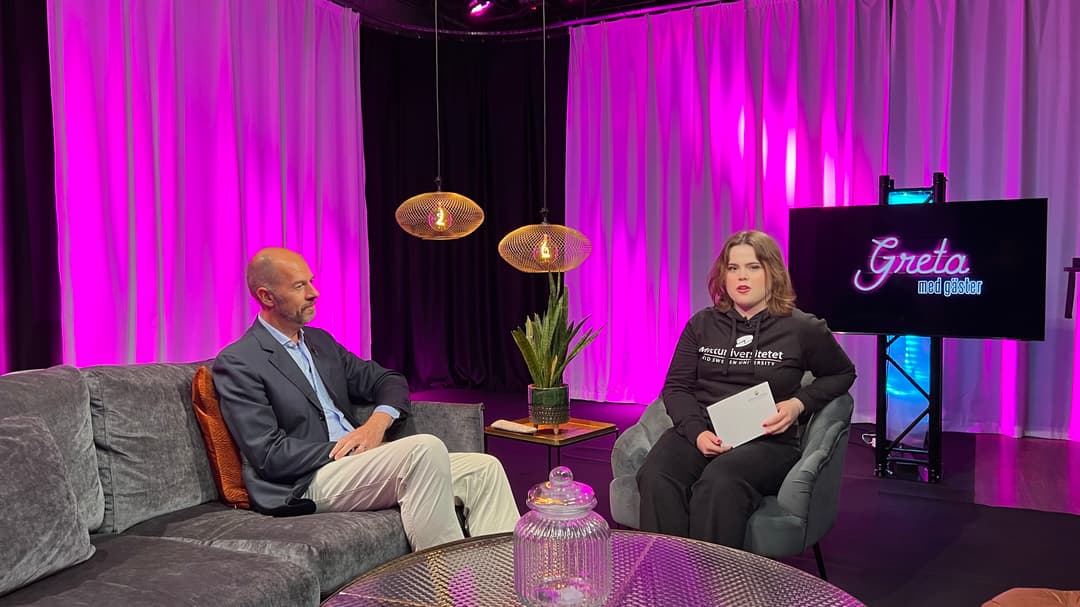 Två personer sitter i en studio, en i en soffa och en i en fåtölj.