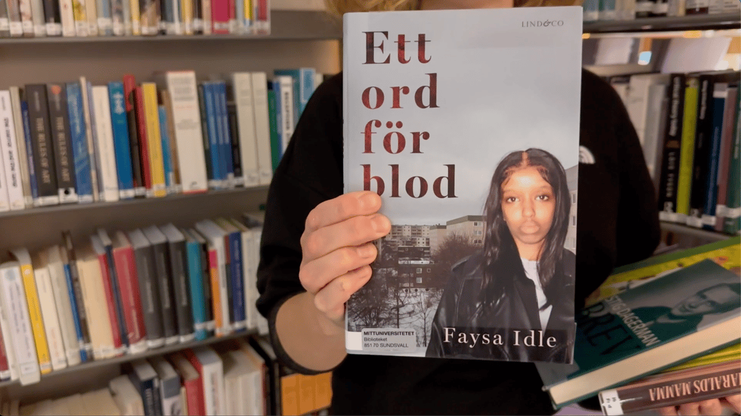 en hand håller upp en bok, bokhyllor i bakgrunden