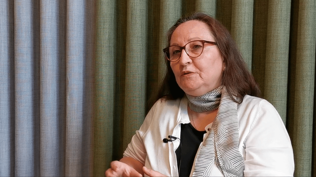 A woman with long brown hair wearing a white blouse and a green scarf is interviewed.