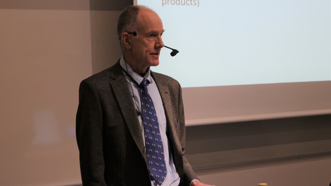 A man in jacket and tie, with a mic standing in front of a whiteboard