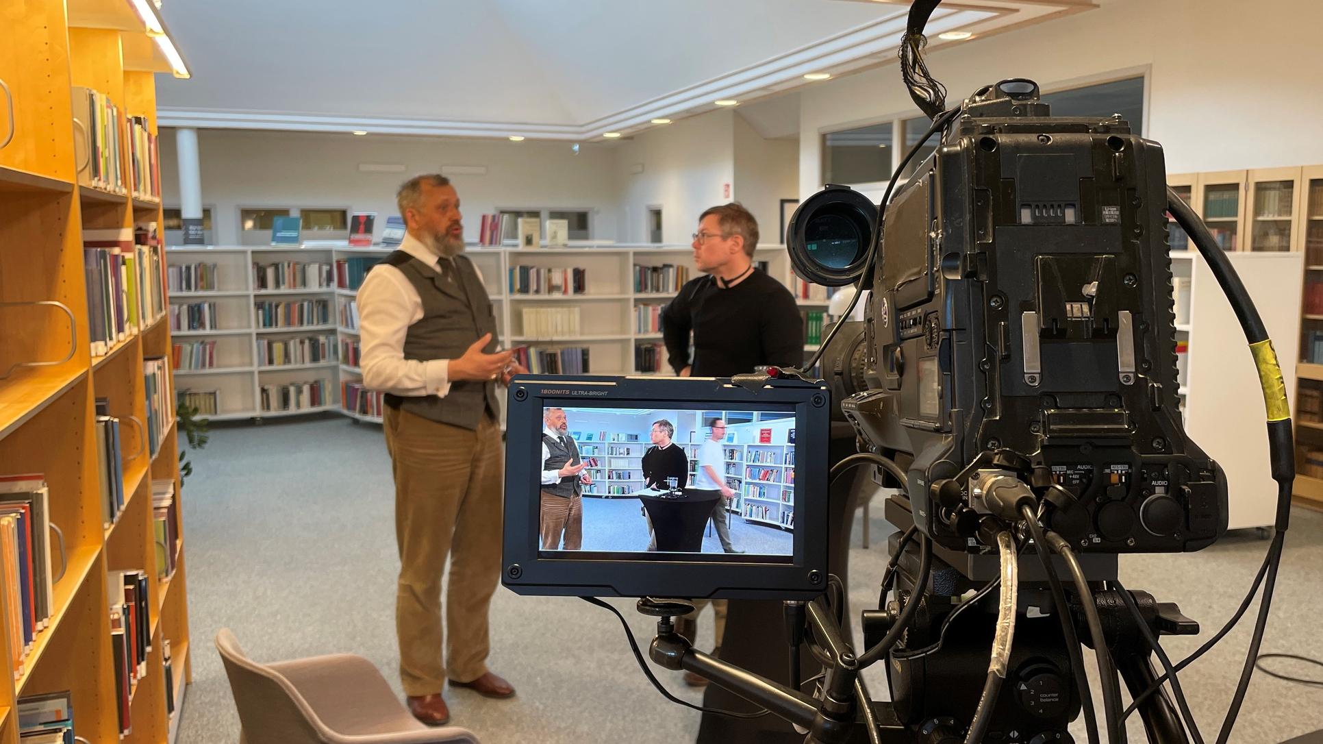 Två män står i ett bibliotek och pratar med varandra. I förgrunden syns en filmkamera. Männen syns även på kamerans skärm.