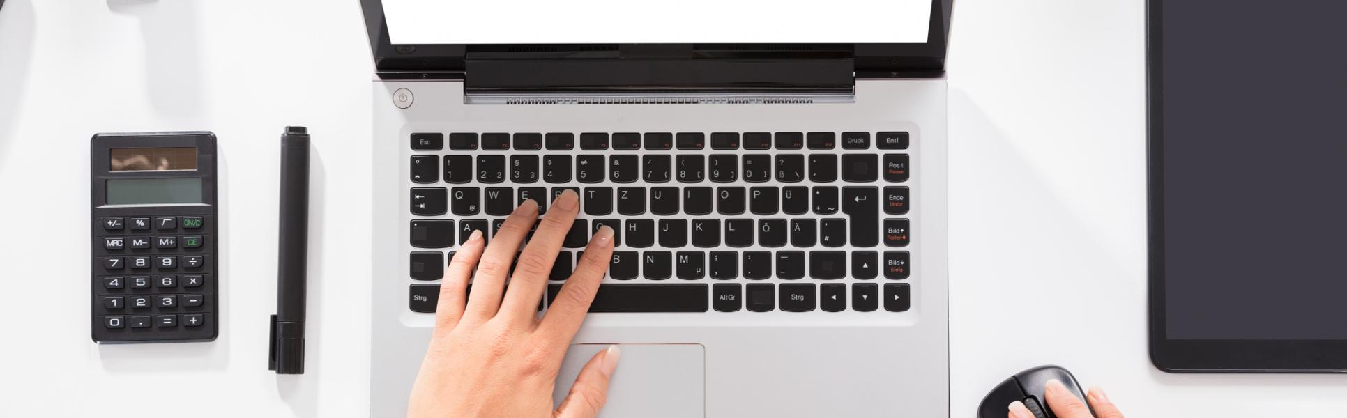 Händer knappar på en laptop, miniräknare, tuschpenna och häftmaskin ligger in till