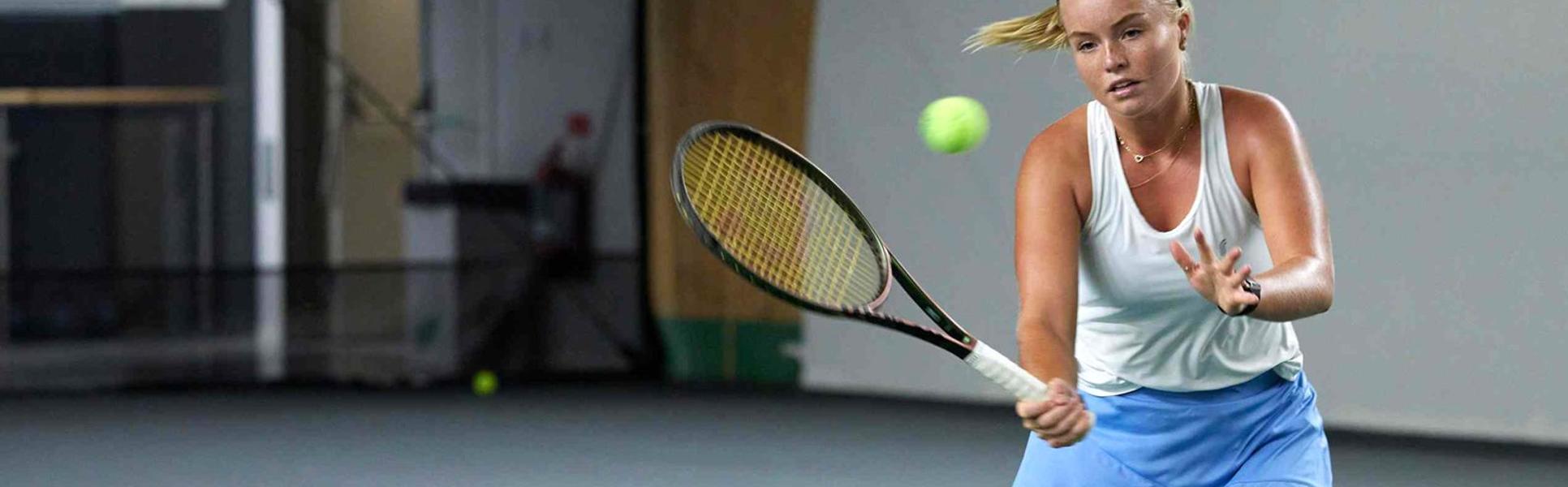 Kvinna som spelar tennis inomhus, hon står vid nät och ska till att slå en volley.