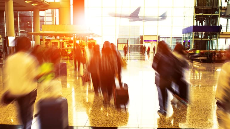 Människor går inomhus på flygplats med resväskor och ett flygplan syns genom fönstret utanför 