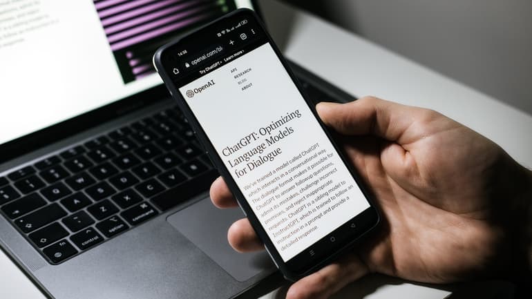 Hand som håller i en mobiltelefon med texten ChatGPT en dator i bakgrunden. 