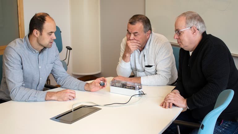 Three men sit at a table and discuss a prototype lying on the table.
