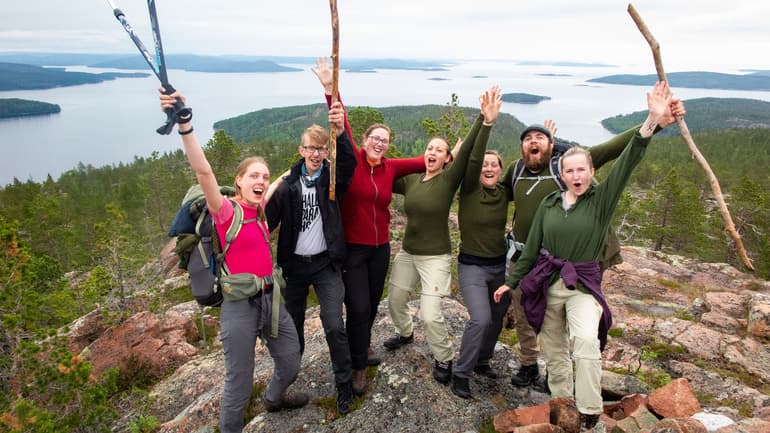 En grupp människor jublar över att ha nått toppen, i bakgrunden utsikt över skog och vatten.