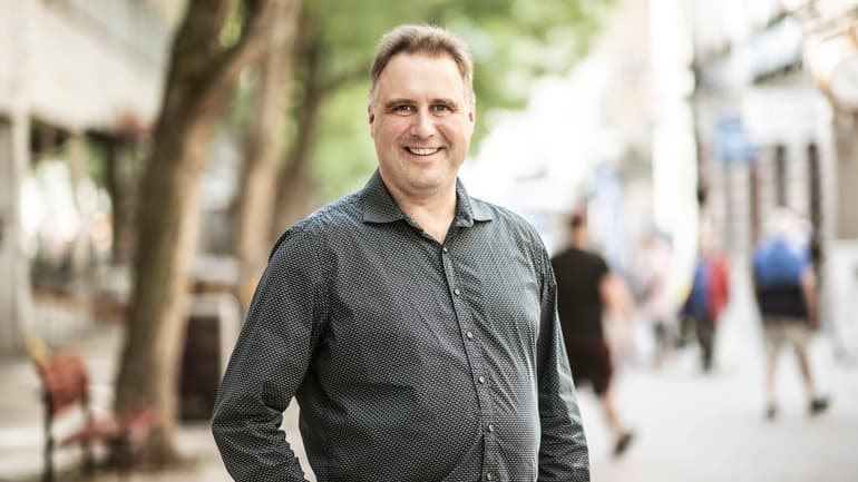 Man med ljusbrunt hår, klädd i en grå skjorta ler mot kameran