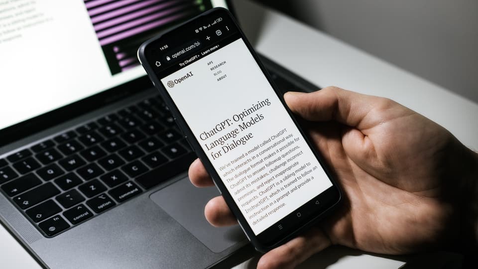Hand som håller i en mobiltelefon med texten ChatGPT en dator i bakgrunden. 