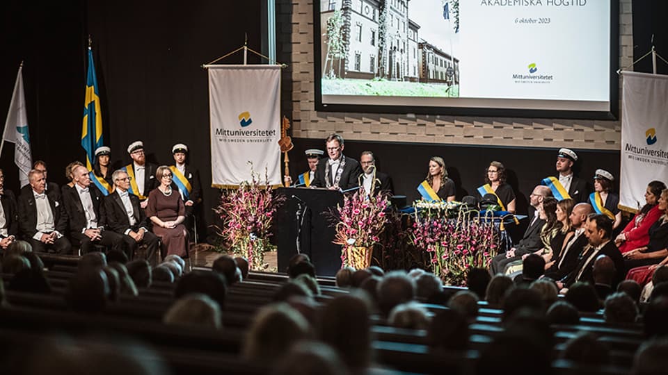 En scen med ceremonideltagare och en man i talarstol. 