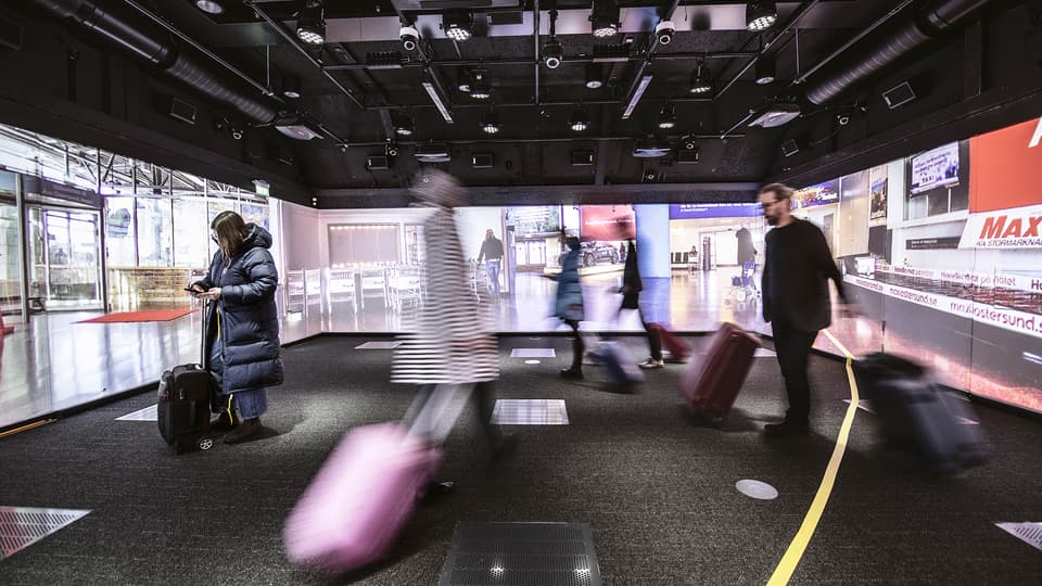 Flera personer står i ett rum med bilder från en flygplats projicerade på väggarna. De har ytterkläder och resväskor. På golvet sitter en gul tejpremsa. I taket finns massor av teknisk utrustning.