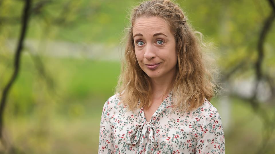 Kvinna med lockigt axellågt hår och blommig blus står utomhus med grönskande träd runt om kring sig.