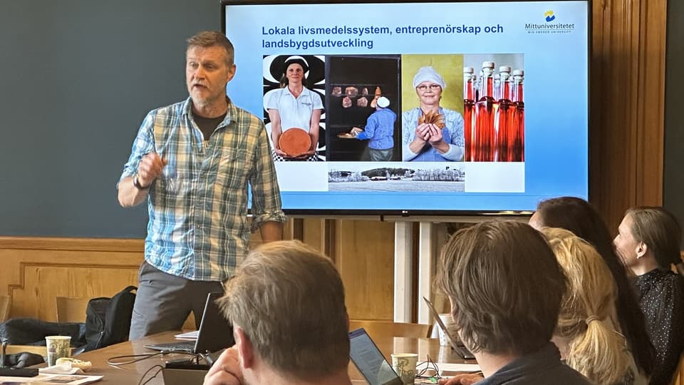 En man med rutig skjorta står framför sittande publik och presenterar med en skärm bakom sig