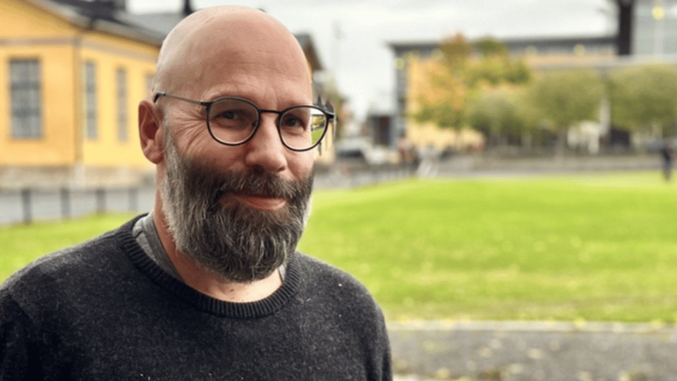 En man med glasögon och en mörkgrå tröja som står utanför framför gula byggnader och ler in i kameran