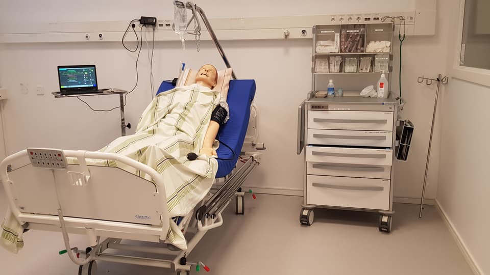 Patientsimulator i en säng i sjukhusmiljö.