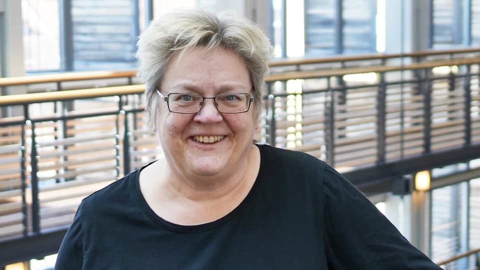 En kvinna står inomhus med fönster och ett räcke bakom sid. Hon har glasögon och en svart tröja. Hon ler. 