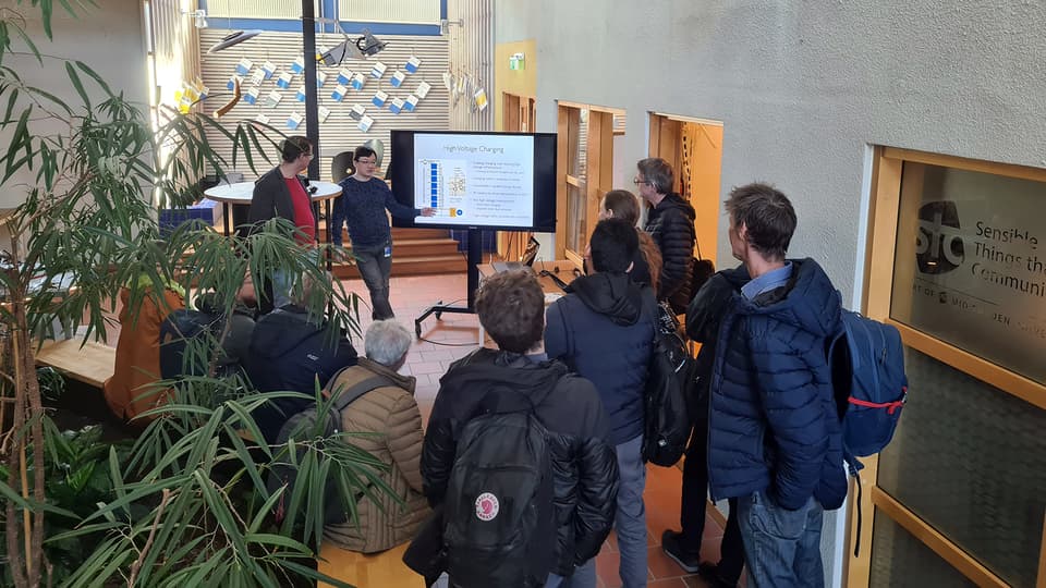 A group of people standing with their backs to the camera watching a television and a man presenting.