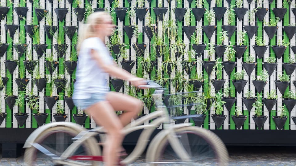 En kvinna med blont hår cyklar och i bakgrunden syns en vägg av växter