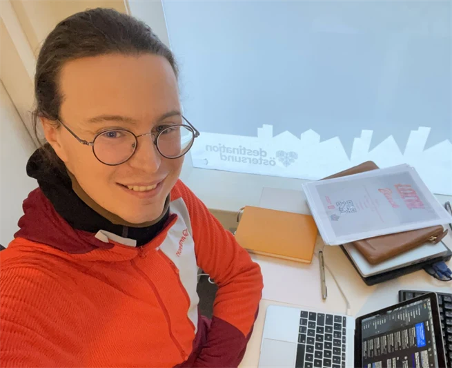 Ung man i glasögon och orange tröja som sitter med sin dator vid ett skrivbord  