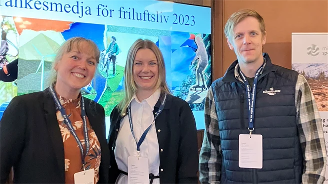 Två kvinnor och en man står i ett rum och tittar in i kameran. Bakom dem finns en skärm med en presentation på och de har namnskyltar hängande runt halsen 