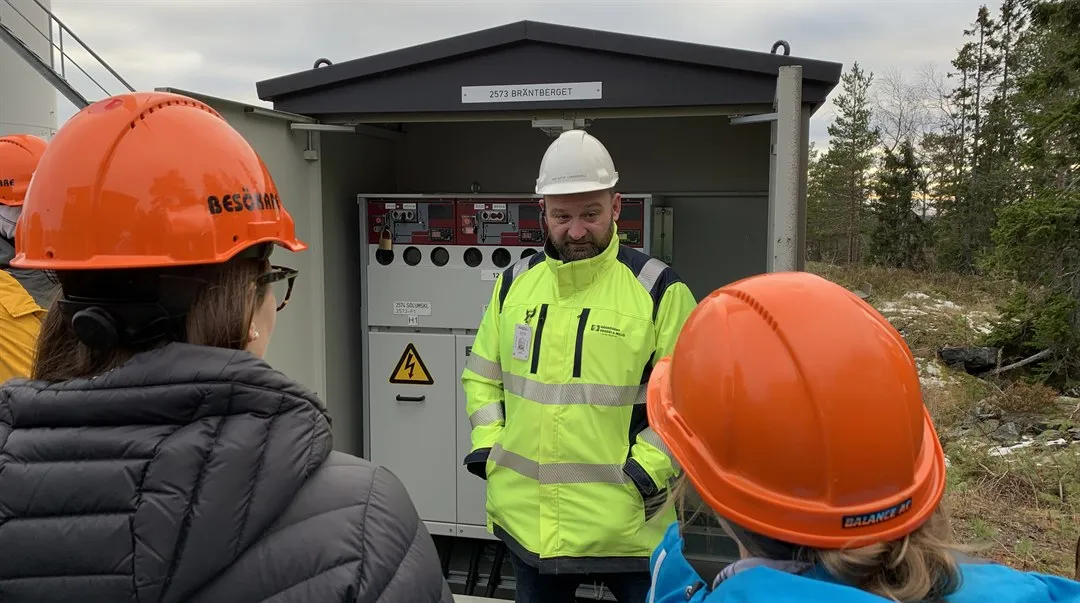 Man i gul varseljacka står framför ett kraftskåp och pratar till människor som har orangea hjälmar.
