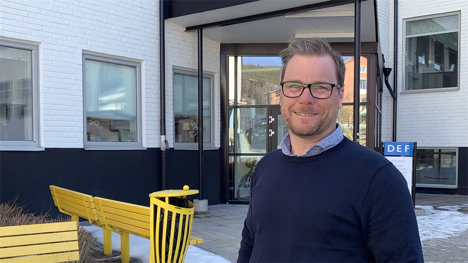 A man in glasses and navy blue sweater standing outside a white house with yellow benches in the background