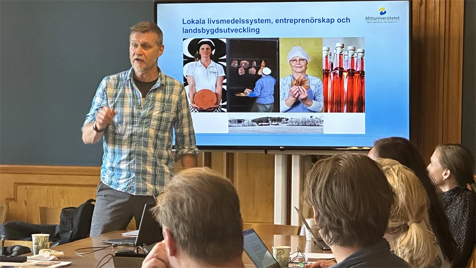 En man med rutig skjorta står framför sittande publik och presenterar med en skärm bakom sig