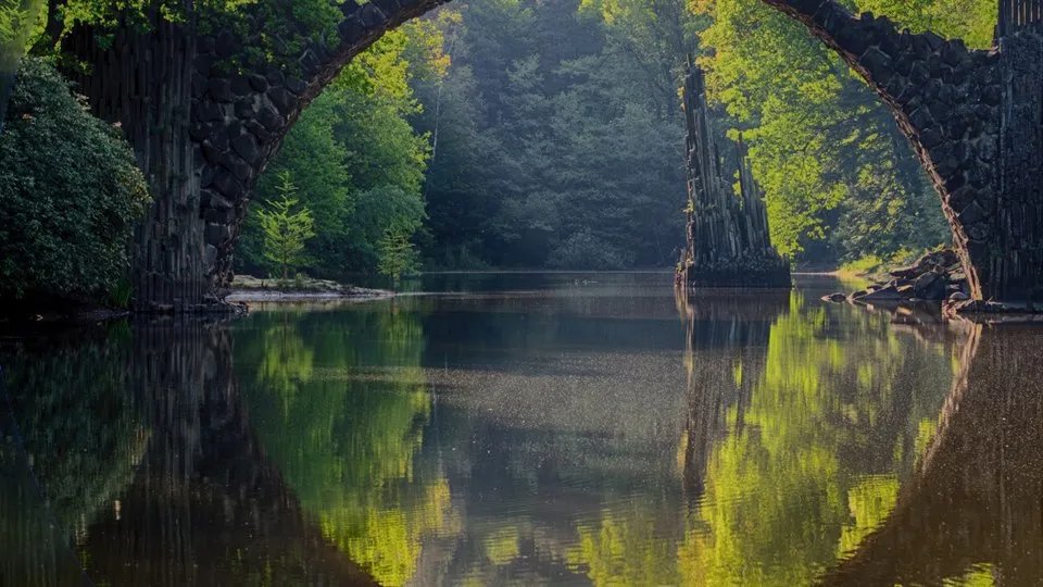 En halvcirkelformad stenbro speglas i vattnet under den. Bron och speglingen skapar en cirkel. I bakgrunden syns lummiga träd.