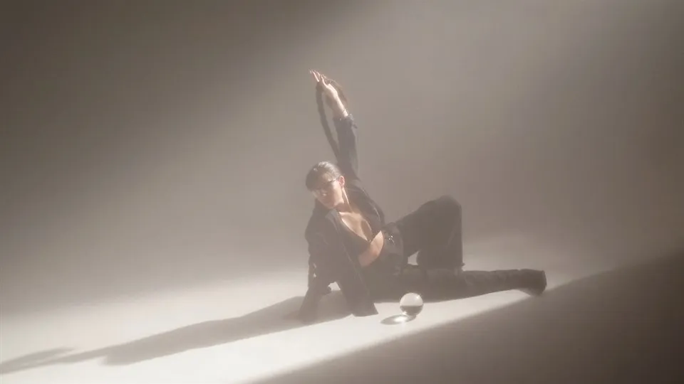 En svartklädd kvinna halvligger på golvet i ett tomt rum med ena armen höjd i luften. Rummet är mörkt och disigt, förutom strålkastarljus på kvinnan.. 