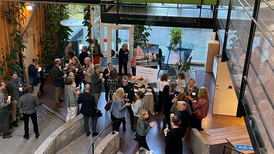 Ett 30-tal människor står i smågrupper och pratar. Många har smörgås eller frukt i handen. Bilden är tagen snett uppifrån och i förgrunden syns ett metallräcke.