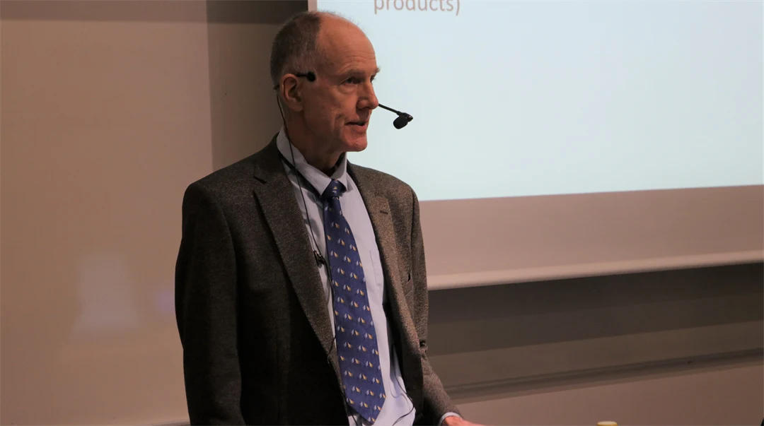 A man in jacket and tie, with a mic standing in front of a whiteboard
