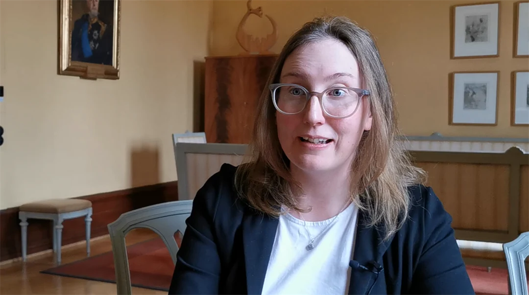 A woman with long brown hair down to her shoulders and wearing a black jacket is interviewed.