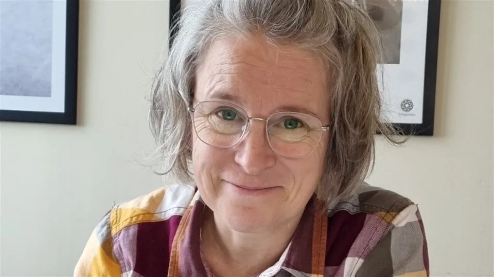 En gråhårig kvinna i glasögon sitter vid ett skrivbord. Hon tittar in i kameran och ler.