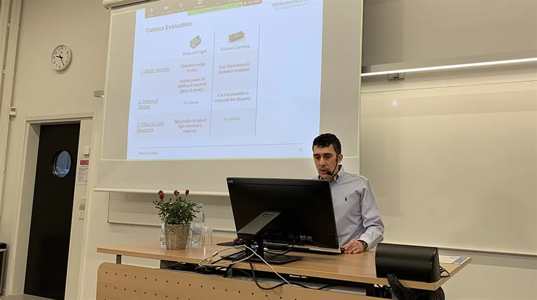 Man standing in front of computer presenting his thesis that can be seen on a screen in the background.