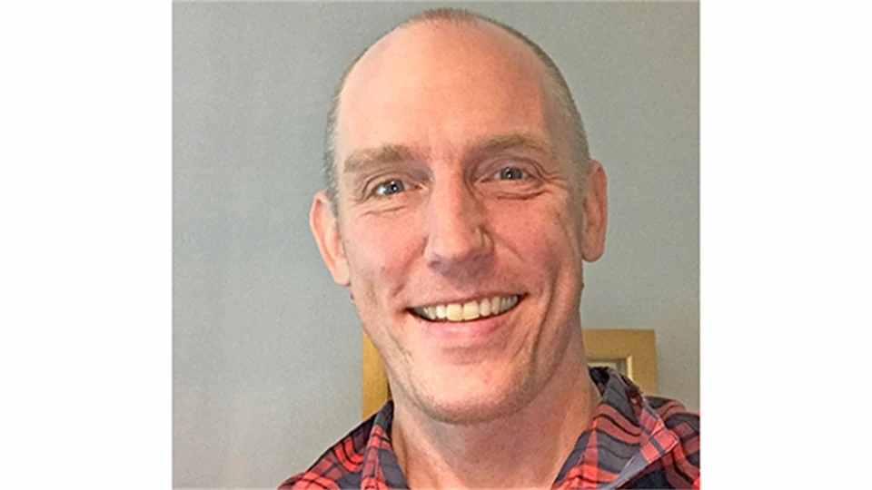 En glad man ler mot kameran klädd i en rödrutig flanellskjorta.