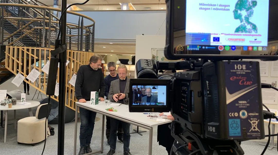 Filmkamera som filmar tre män som samtalar vid ett bord.