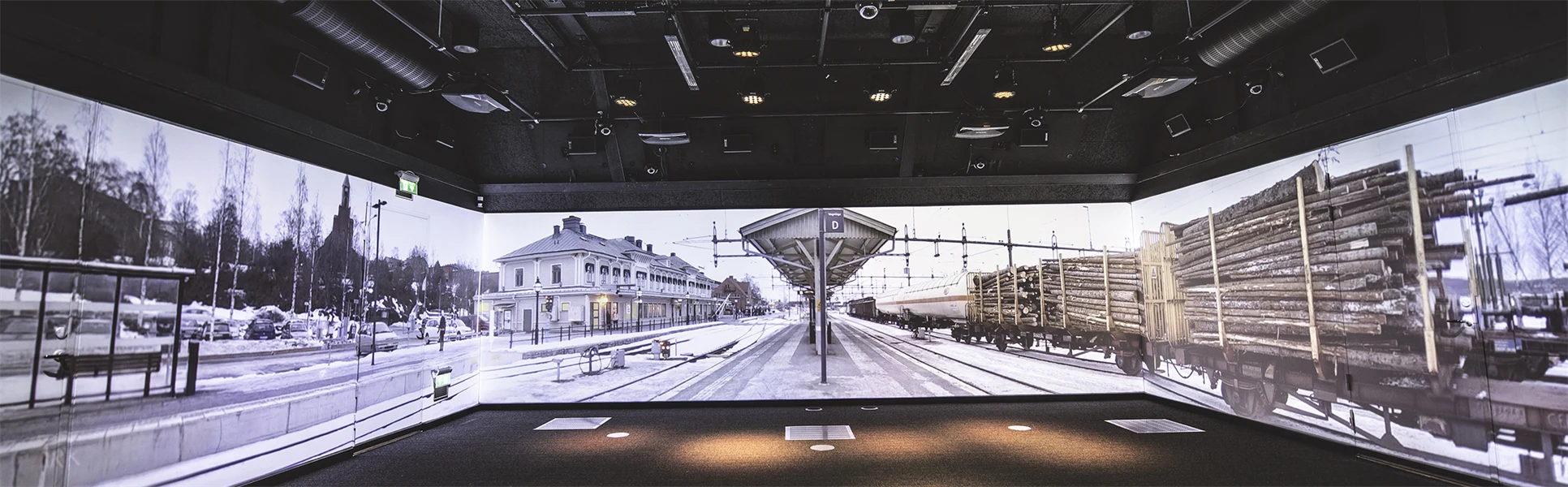 A large room with black floor and ceiling. Images from a railway platform are projected on the walls. There are lots of technical equipment in the ceiling.