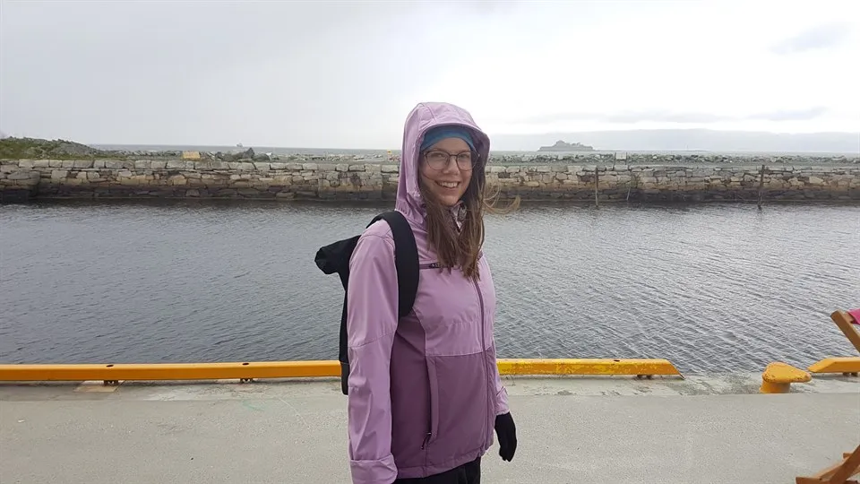 En kvinna i lila regnjacka och ryggsäck står på en kaj. Hon ler och tittar in i kameran. Bakom henne ser man vatten och en stenpir.