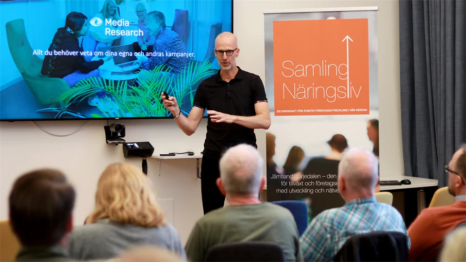 A man standing next to a large screen and speaking to a seated audience
