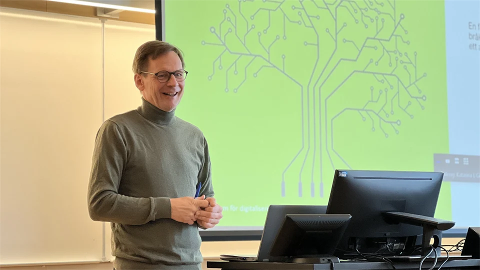 En man med grå tröja står och glasögon pratar framför en projektorduk