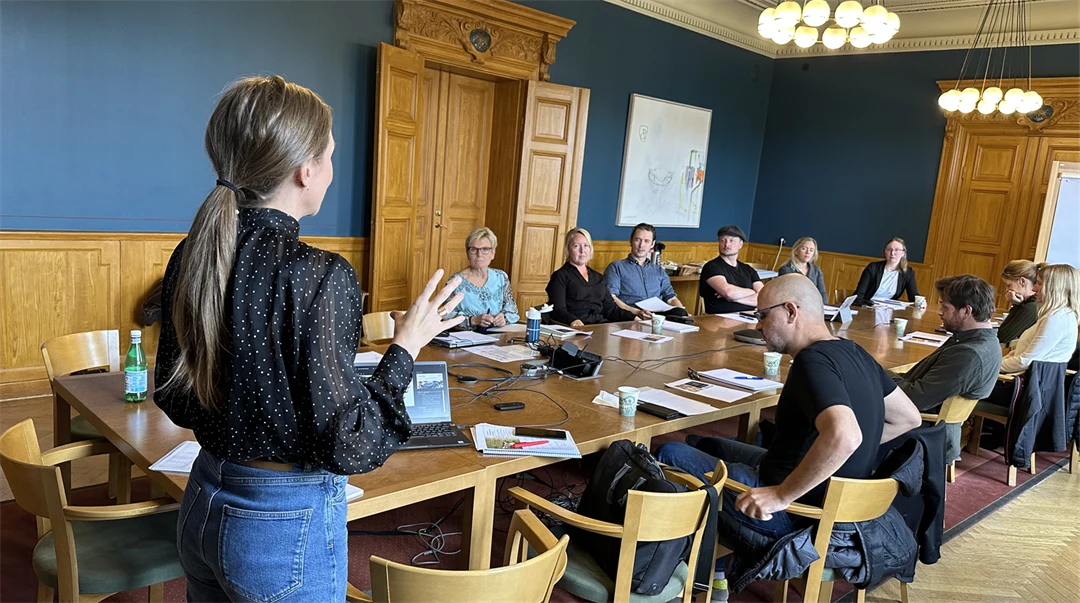 Kvinna står upp framför ett tiotal personer som sitter vid ett långt bord. Hon gestikulerar med händerna.