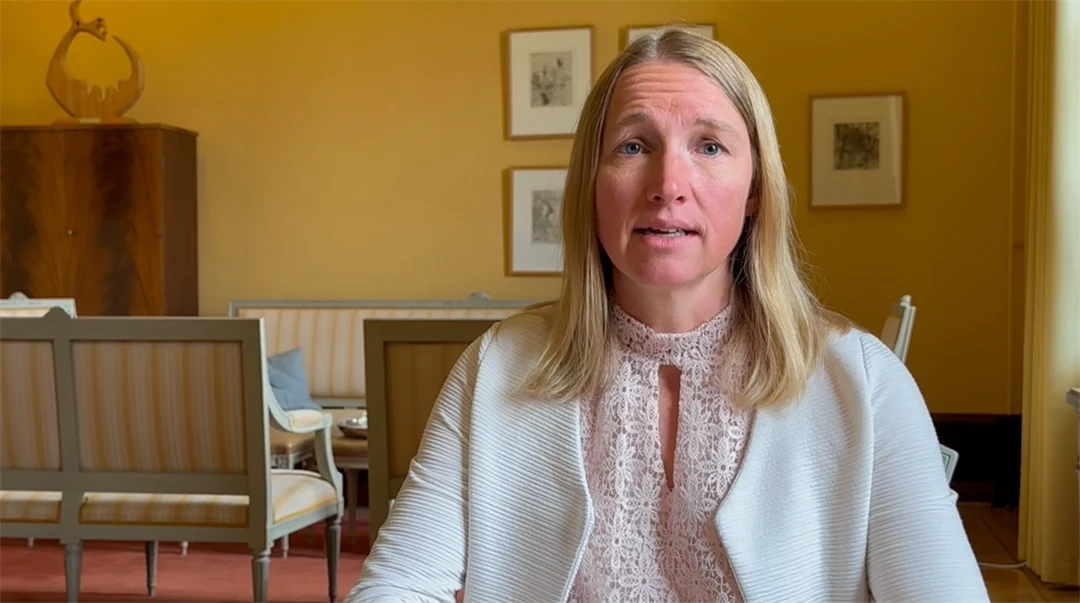 A blonde woman in a white jacket sits in a room with yellow walls and a seating area in the background. Paintings hang on the walls. 