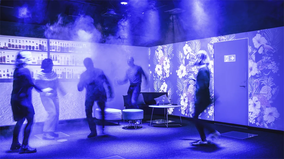 Four people dance in a room with dim lighting. On the walls, a nightclub environment with a bar and an emergency exit is projected. In one corner are some poufs and a low table.