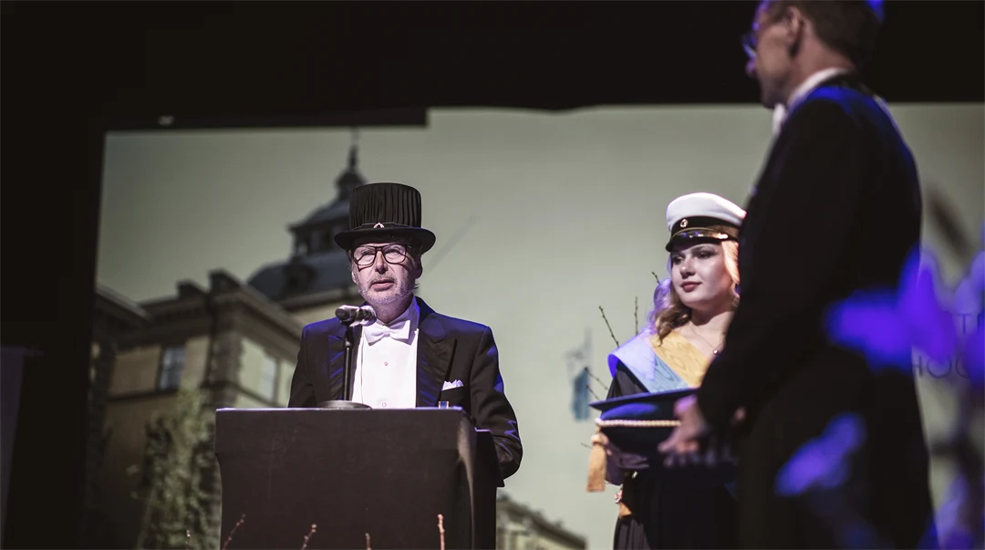 A well-dressed person with a doctor's hat stands in a lectern.