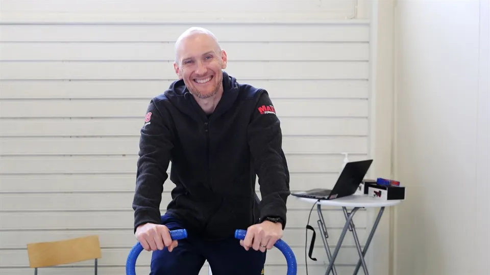 En leende mann sitter på en testcykel, bakom står ett bord med en bärbar dator.