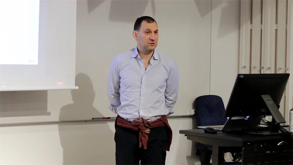 A man with dark hair and a light shirt stands in front of a whiteboard