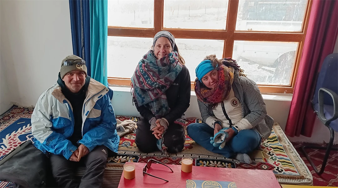 Tre personer sitter på en matta inomhus. De har jackor, mössor och stora halsdukar på sig.