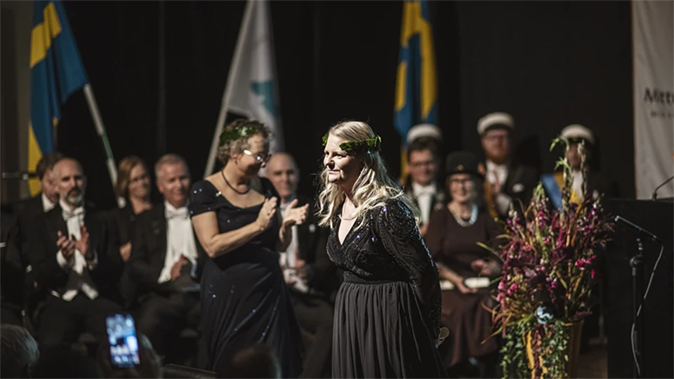 A well-dressed woman with a laurel wreath stands on a stage. 
